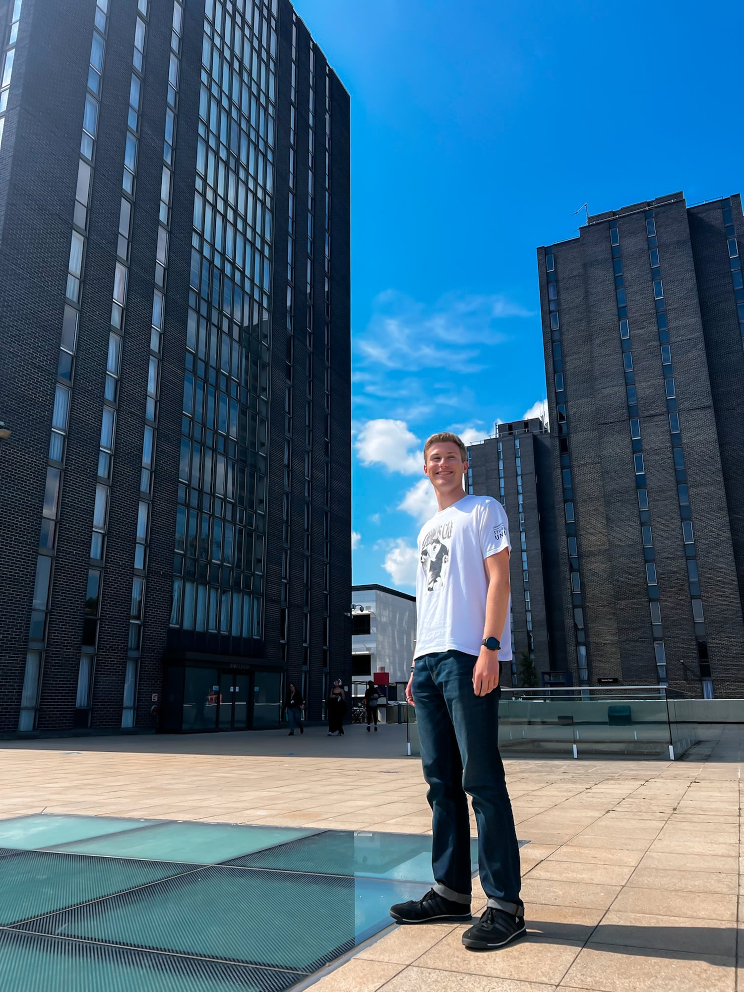 A young man stands outside in front of two large skyscraper buildings and a blue sky, he wears a white t-shirt and jeans. 