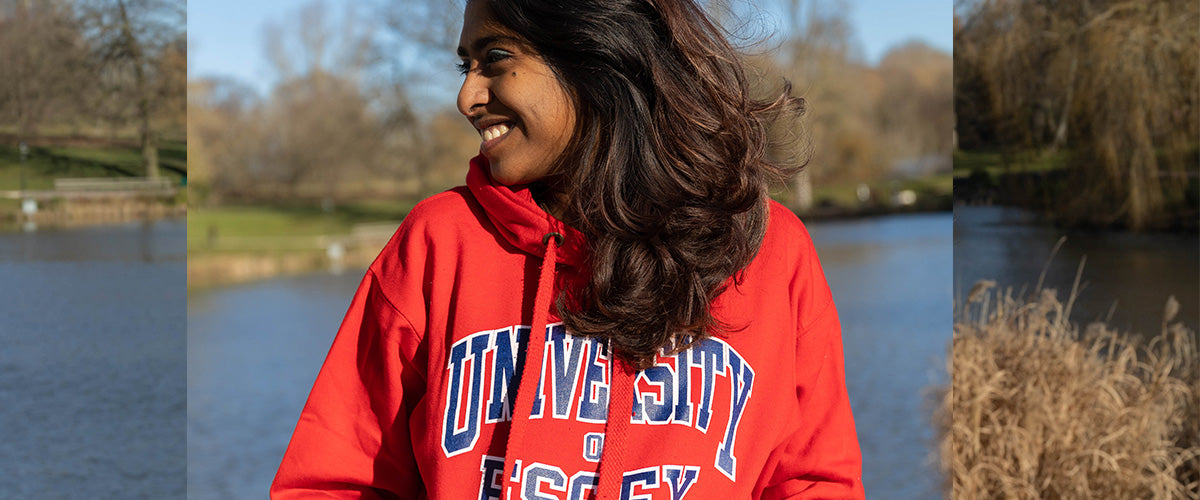 A young woman wears a red hoodie with "University of Essex" in varsity style font, there is a pond and foliage behind her